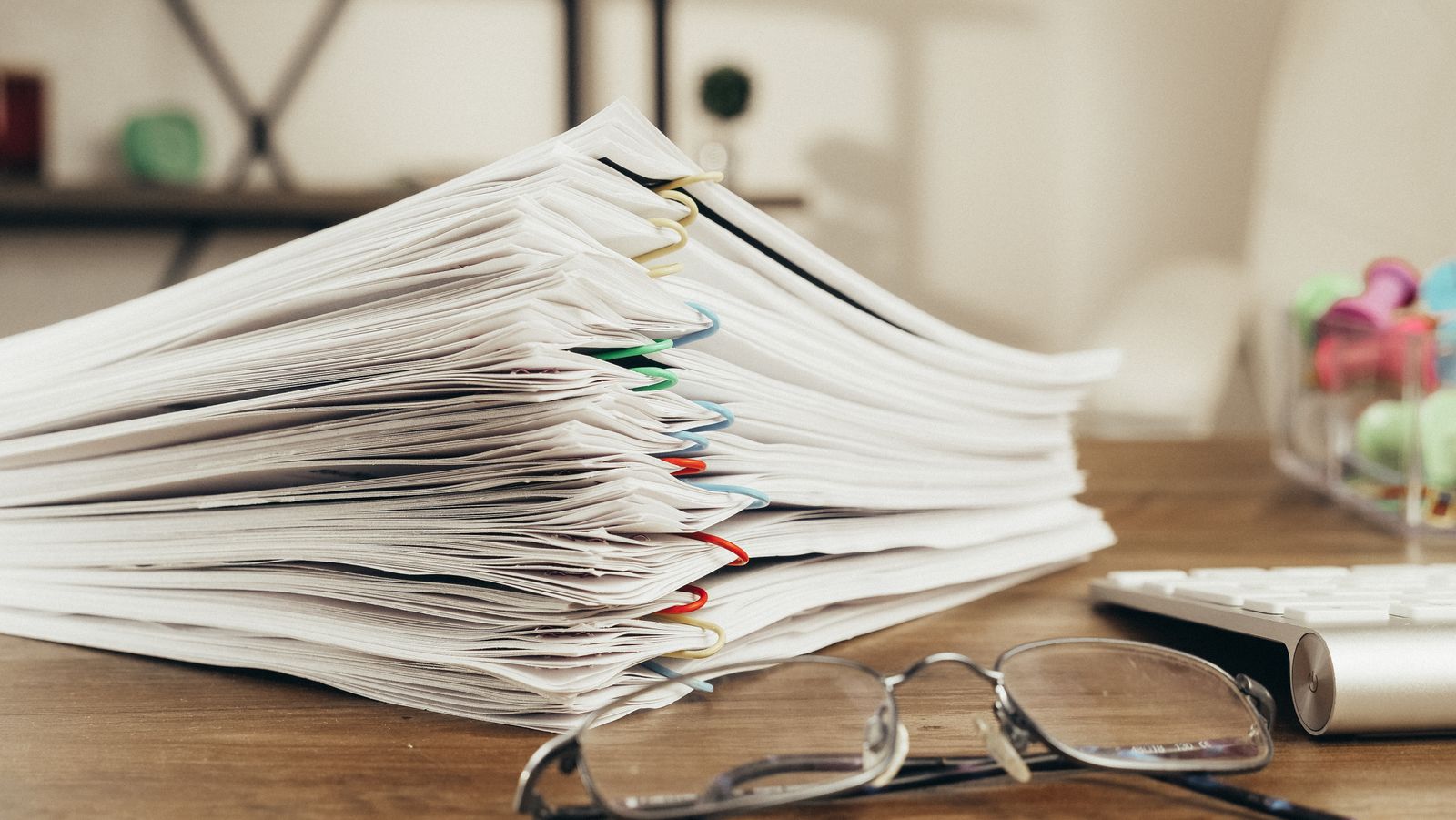 A stack of clipped documents on a desk beside a pair of reading glasses
