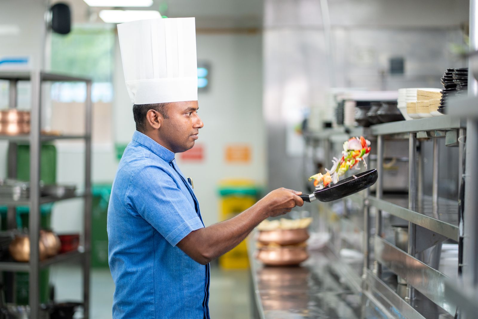 A chef in a toque cooking at a stainless-steel station in a commercial kitchen